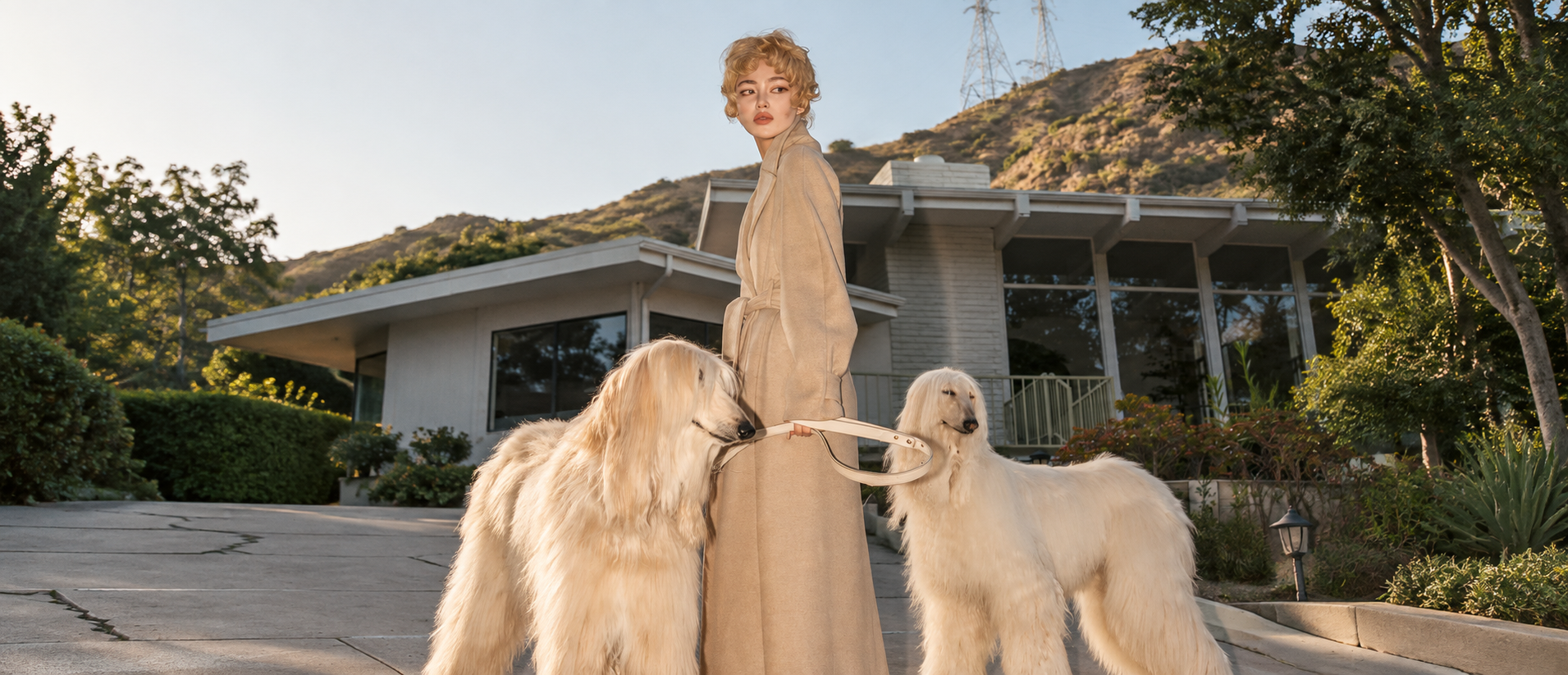 Una mujer de tapado camel posa frente a una casa moderna con sus dos lebreles afganos de pelo claro