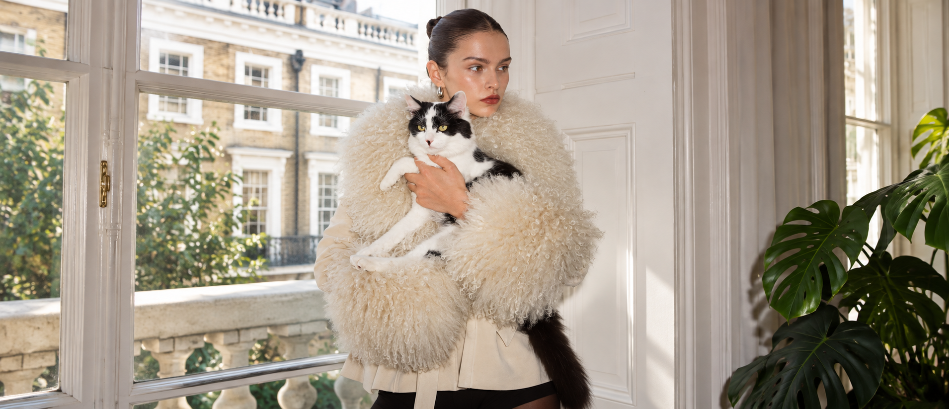 Una mujer con un tapado de piel clara sostiene en brazos a su gato blanco y negro, junto a una ventana