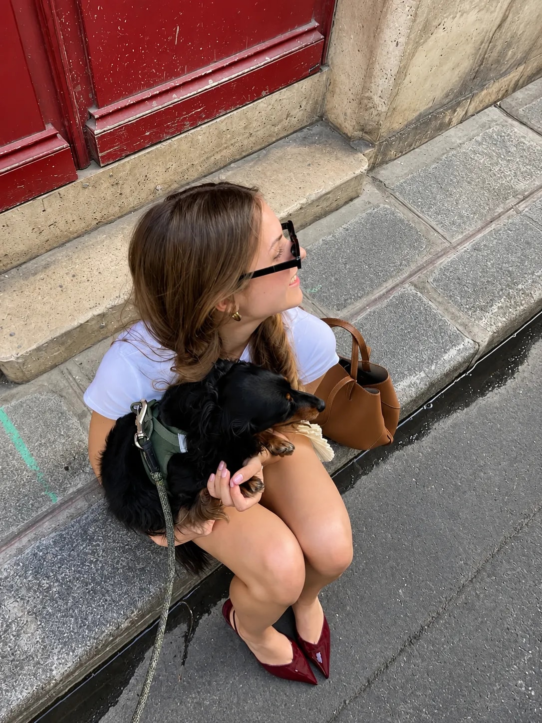 Una chica sentada en el escalón de una puerta roja abraza a su perro salchicha negro, con pretal verde y correa